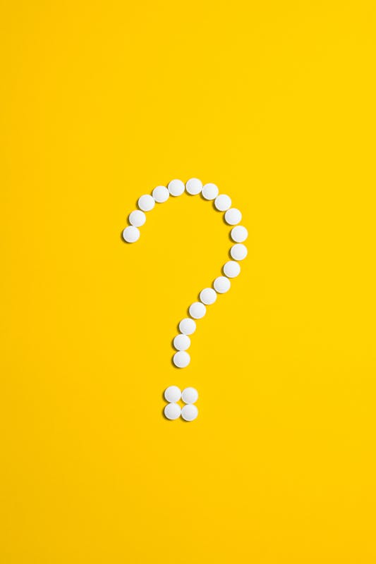 White pills arranged in a question mark shape on a bright yellow background, symbolizing medical queries.