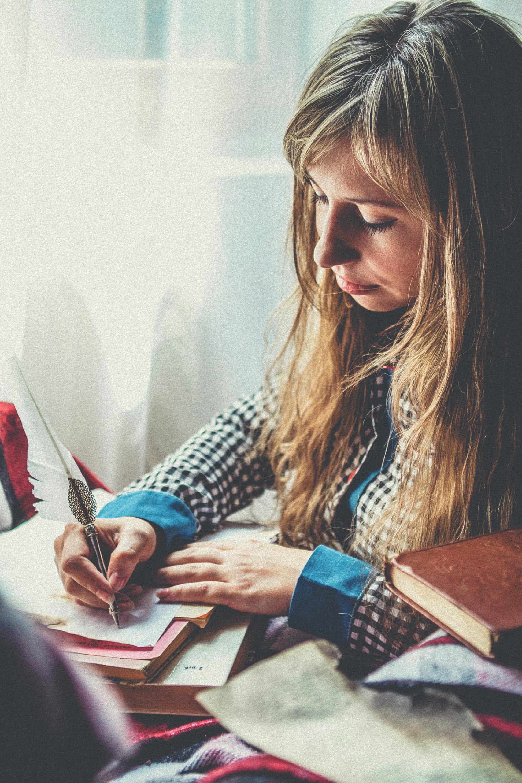 Woman with blonde hair writing in a notebook by a window, creating a thoughtful and serene atmosphere.