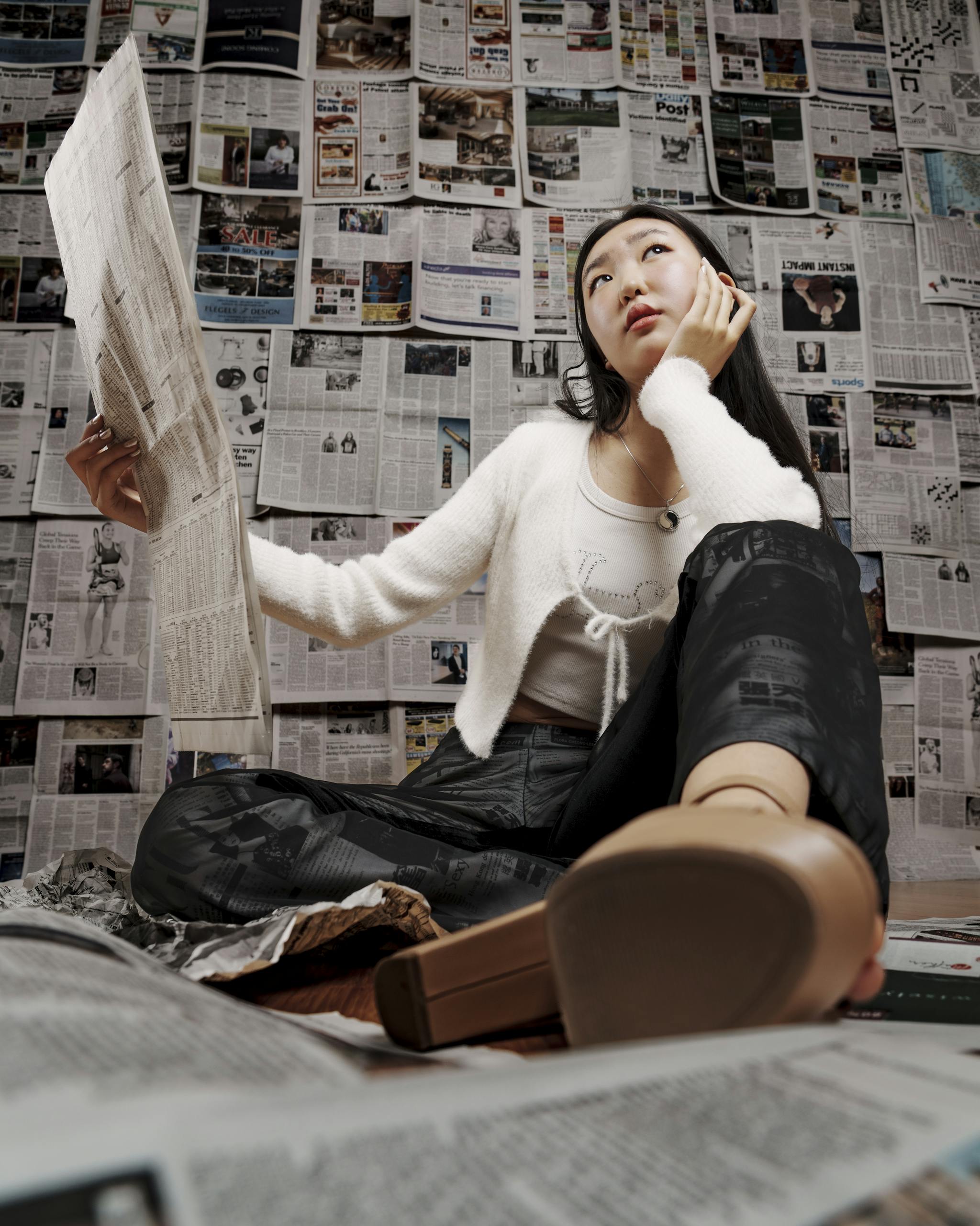 Young woman sitting and thoughtfully reading a newspaper indoors, surrounded by news articles.